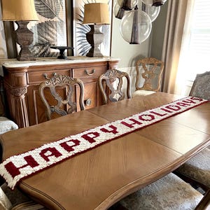 May include: A wooden dining table with a cream and burgundy "HAPPY HOLIDAYS" table runner. The table is surrounded by ornate chairs. Two lamps with tan shades sit on a wooden cabinet, with framed botanical prints in the background.