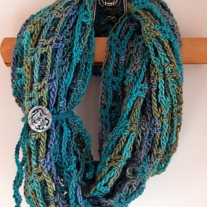 May include: A handmade infinity scarf in shades of teal, blue, and olive green, with a decorative silver button closure. The scarf is made of a chunky knit yarn and is displayed on a wooden hanger.