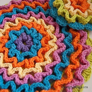 May include: A colourful crocheted doily with a scalloped edge. The doily is made in shades of pink, orange, yellow, blue and green.