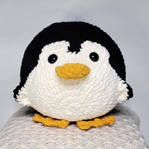 May include: A handmade, plush penguin toy. The penguin has a white body, black head and wings, and a yellow beak and feet. The eyes are black. The toy is made of a soft, textured material.