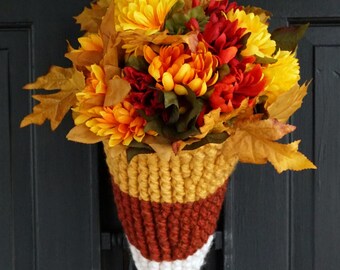 Pode incluir: Um pendente de parede em forma de cone com uma capa de lã tricotada à mão em riscas horizontais amarelas, cor de laranja e brancas. O topo está preenchido com um arranjo denso de flores e folhas artificiais em tons de dourado, vermelho e castanho.
