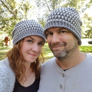 May include: Two people wearing matching gray and white crocheted beanies. The beanies have a textured pattern. The background is a blurred outdoor scene with trees and sunlight. The people are smiling at the camera.