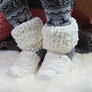 May include: A pair of plush, white and gray crocheted slippers featuring a thick, textured cuff. The slippers are worn over gray and black patterned leggings that display a repeating design of reindeer and snowflakes in a winter theme.