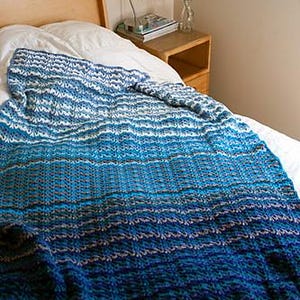 May include: A rectangular crochet blanket featuring a gradient color pattern that transitions from light blue and white at the top to deep navy blue at the bottom. The textile displays a repeating textured stitch pattern throughout the entire surface.