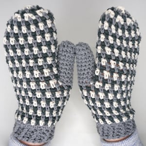 May include: A pair of handmade mittens in a checkerboard pattern. The mittens are primarily gray, with a pattern of white and dark gray squares. The cuffs and thumbs are solid gray. The mittens are designed for warmth and comfort.