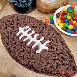 May include: A brown crocheted football with white stitching. The football is on a wooden surface.