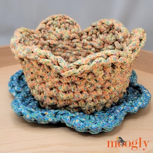 May include: A handmade crochet basket with a floral pattern in yellow, orange, and green, sitting on a blue floral coaster. The basket has a scalloped edge and is made of thick, soft fabric. The coaster is also crocheted.