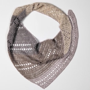 May include: A handmade, triangular crochet shawl in shades of grey, brown, and beige. The shawl features an open, lacy stitch pattern with a gradient effect, transitioning from darker to lighter tones. The shawl is displayed on a white background.