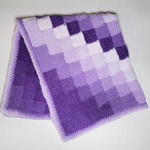 Può includere: Una copertina per neonati in crochet di colore viola e bianco con un motivo a scacchi. La copertina è realizzata con filato morbido e ha una texture soffice.