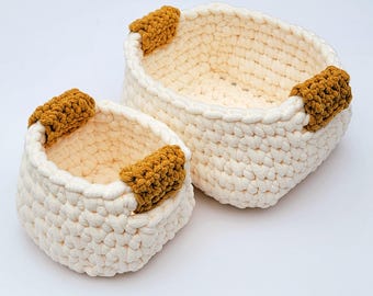 May include: Two off-white crochet baskets of different sizes, each featuring two mustard yellow textured handles attached to the sides. The baskets are constructed from thick yarn with a visible knotted stitch pattern and an open top design.