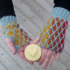 May include: Crocheted fingerless gloves in shades of blue, yellow, orange, and red. The gloves feature a lattice design and are held by a person. A round, light-colored cookie with a heart shape is held between the gloves.