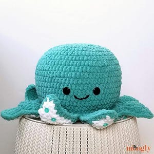 May include: A teal crocheted octopus plush toy with white accents and black eyes. The octopus is sitting on a white wicker stool.