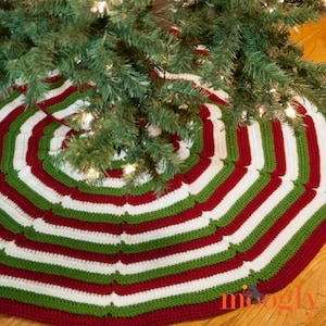 May include: A handmade Christmas tree skirt with a striped pattern in red, green, and white. The skirt is crocheted and has a scalloped edge. The tree skirt is placed under a green Christmas tree.