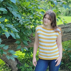 May include: A yellow and white striped crochet top with a round neck. The sleeveless top is worn with blue jeans. The image is set against a backdrop of green leaves and a tree trunk.