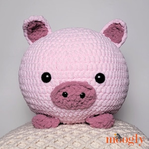 Puede incluir: Un juguete de peluche de cerdo rosa de crochet con ojos negros y un hocico rosa. El cerdo está sentado en una manta de punto blanca.