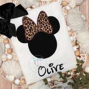 May include: White t-shirt featuring a black Minnie Mouse design with a leopard print bow. The name "Olive" is written in black script below the design. A large black bow is visible in the upper left corner.