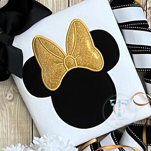 May include: A white fabric item featuring a black mouse head design with a gold glitter bow. The bow is outlined in gold. A black bow and black and white striped ribbon are visible in the background.