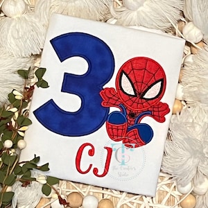 May include: White t-shirt featuring a large blue number 3 and a cartoon Spider-Man graphic. The Spider-Man is red and blue. The shirt also has the initials "CJ" in red.