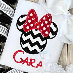 May include: White t-shirt featuring a Minnie Mouse design. The design includes a red polka-dot bow, black and white chevron ears, and the name "Gara" in red. The shirt is decorated with a bow and ribbon.