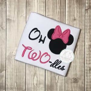 May include: White t-shirt with the text "Oh Two-dles" in black and pink script. A black mouse head with a pink polka dot bow is on the right side. The shirt has a round logo in the bottom right corner.