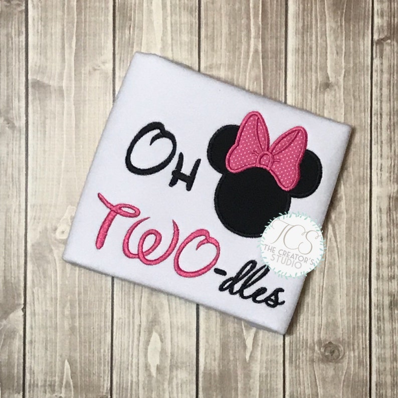 May include: White t-shirt with the text "Oh Two-dles" in black and pink script. A black mouse head with a pink polka dot bow is on the right side. The shirt has a round logo in the bottom right corner.