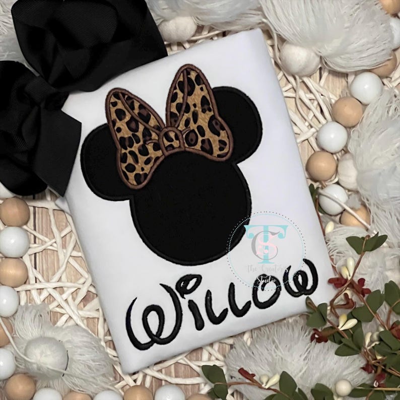 May include: White t-shirt featuring a black Minnie Mouse design with a leopard print bow. The name "Willow" is written in black script below the design. A large black bow is visible in the upper left corner.