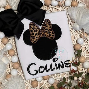 May include: White t-shirt featuring a black Minnie Mouse design with a leopard print bow and the name "Collins" in black script. A large black bow is placed on the top left. The shirt is surrounded by decorative beads and greenery.