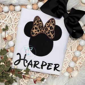 May include: White t-shirt featuring a black Minnie Mouse design with a leopard print bow and the name "Harper" in black script. A large black bow sits at the top right. The shirt is likely for a child.