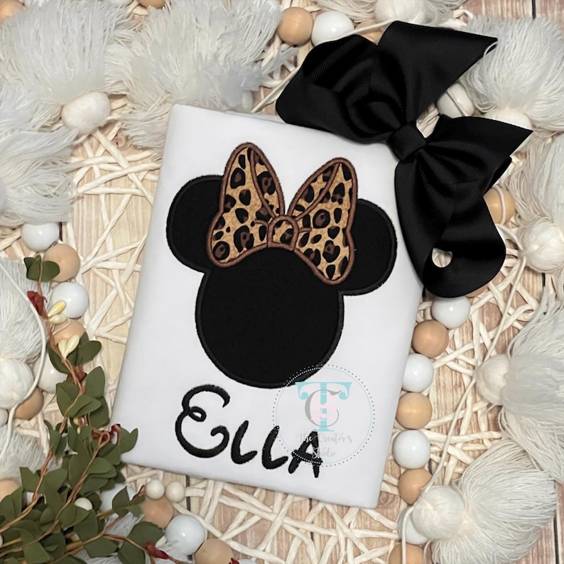 May include: A white shirt featuring an embroidered black silhouette of a mouse head with a leopard print bow. The name Ella is embroidered in black script below the design. A large black hair bow is attached to the top right of the shirt.