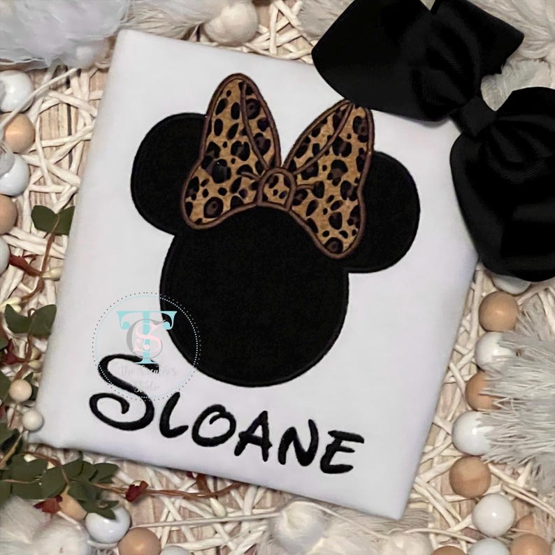 May include: White t-shirt featuring a black Minnie Mouse design with a leopard print bow. The name "Sloane" is embroidered in black script below the graphic. A large black bow is visible in the upper right corner.