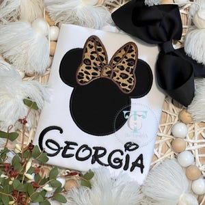 May include: White t-shirt with a black Minnie Mouse design, featuring a leopard print bow and the name "Georgia" in black script. A large black bow is placed on the right. The shirt is surrounded by decorative elements.