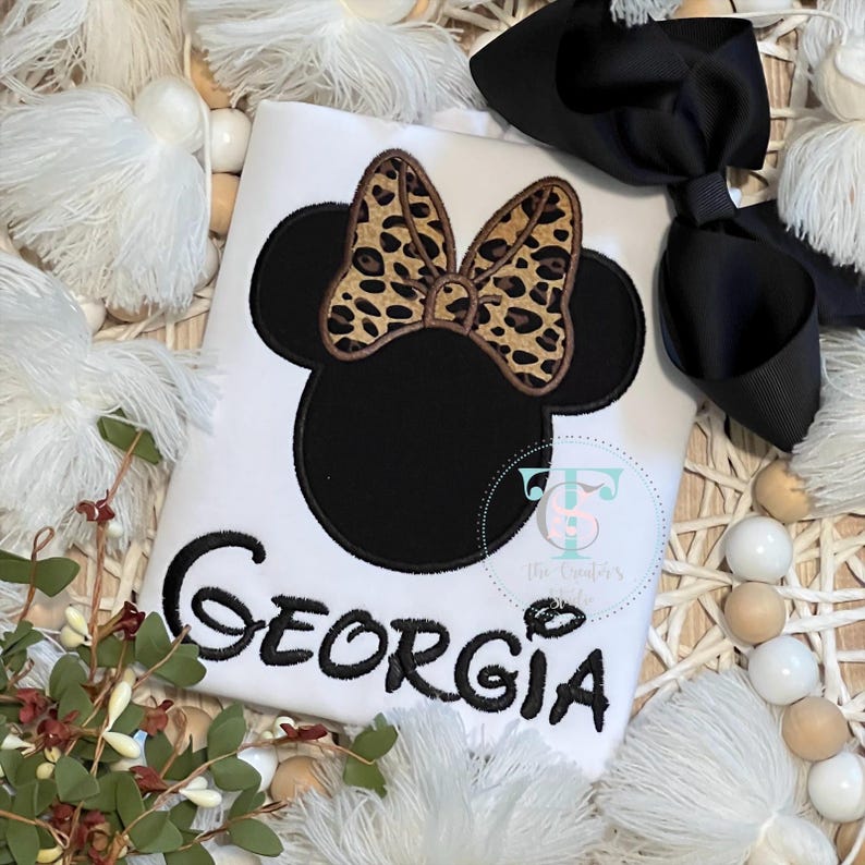 May include: White t-shirt with a black Minnie Mouse design, featuring a leopard print bow and the name "Georgia" in black script. A large black bow is placed on the right. The shirt is surrounded by decorative elements.