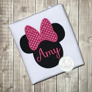 May include: White t-shirt featuring a black Minnie Mouse head design with a pink polka dot bow and the name "Amy" in pink script. The design is embroidered on the shirt. The Creator's Studio logo is in the bottom right corner.