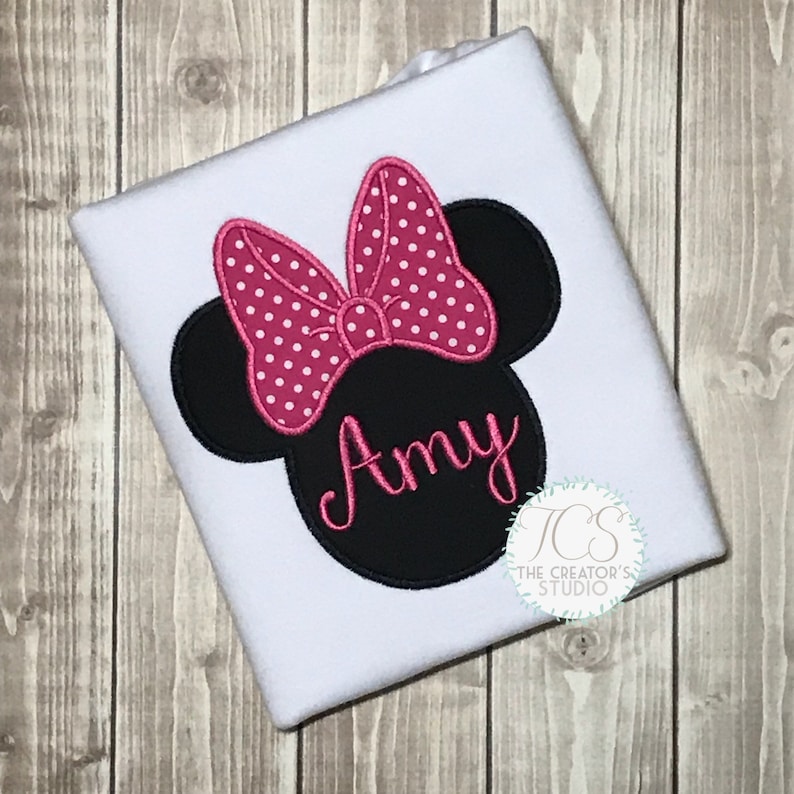 May include: White t-shirt featuring a black Minnie Mouse head design with a pink polka dot bow and the name "Amy" in pink script. The design is embroidered on the shirt. The Creator's Studio logo is in the bottom right corner.