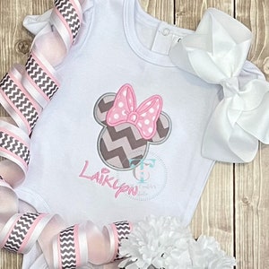 May include: White baby bodysuit with a Minnie Mouse design. The design features a pink polka dot bow and a gray and white chevron pattern. The name "Laiklyn" is embroidered in pink. A white bow and pink and gray ribbon surround the bodysuit.