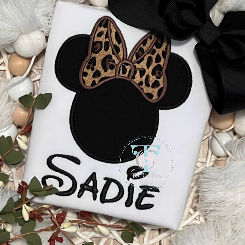 May include: White t-shirt featuring a black Minnie Mouse design with a leopard print bow. The name "Sadie" is embroidered in black script. A black bow is visible in the background.