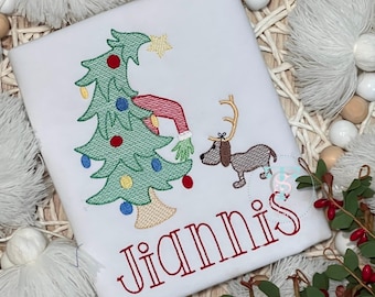 May include: A white t-shirt featuring an embroidered design of a green Christmas tree with colorful ornaments, a red sleeve with a green hand, and a brown dog wearing gold reindeer antlers. The name JIANNIS is embroidered in red below the design.