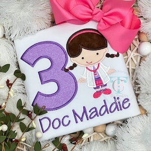 May include: A white shirt featuring an embroidered purple number three and the text Doc Maddie in purple. An embroidered cartoon character wearing a white lab coat, pink skirt, and stethoscope is present. A large bright pink bow is attached.