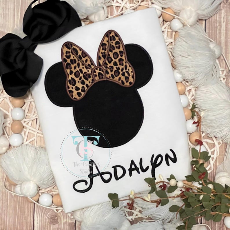 May include: White t-shirt featuring a black Minnie Mouse design with a leopard print bow. The name "Adalyn" is written in black script below the design. A black bow is placed on the top left. The shirt is on a wooden surface.