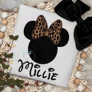 May include: White t-shirt featuring a black Minnie Mouse design with a leopard print bow and the name "Millie" in cursive. A large black bow is also visible. The shirt is likely for a child.