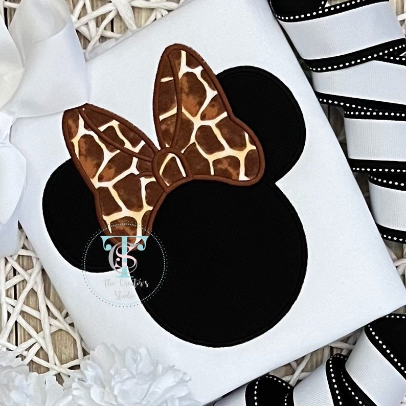 May include: White t-shirt featuring a black Mickey Mouse head design with a giraffe print bow. The design includes the text "The Creator's Studio" in a circular logo. White and black ribbon is visible in the background.