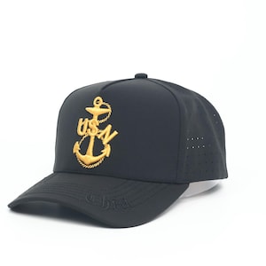 May include: A black baseball cap featuring a gold embroidered anchor emblem with the letters USN centered on the front panel. The cap has a structured crown, a curved brim, and perforated side panels for ventilation.