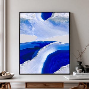 Puede incluir: Una impresión de arte mural enmarcada que presenta una composición abstracta de patrones de piedra ágata en capas azules y blancas con acentos plateados brillantes. El diseño muestra bandas de azul cobalto y azul claro.