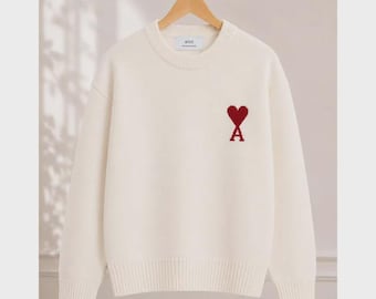 May include: A cream-colored long-sleeve crewneck sweater featuring a red heart and letter A logo embroidered on the left chest. The garment has a ribbed neckline, cuffs, and hem, with an internal tag displaying the text ami.