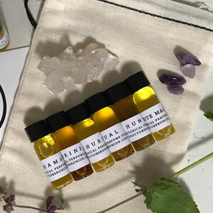 May include: A set of seven small glass bottles with labels containing the names of different essential oils. The bottles are arranged in a circle on a white surface. The labels are black and white and include the words "Botanical Perfume", "Organic", "Local", and "Reunion".