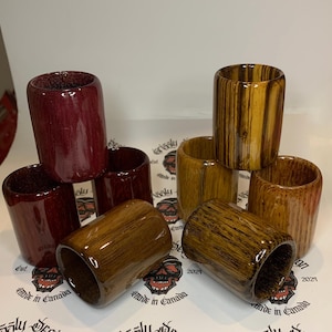 May include: A collection of handcrafted wooden shot glasses in various wood grains and colors. The shot glasses are cylindrical with a glossy finish. Some are a deep red, others a warm brown, and some have a striped pattern.