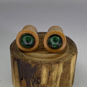 Puede incluir: Par de medidores de oreja de madera con incrustaciones de piedras preciosas verdes y rosas. Los medidores se muestran en un tronco de madera.