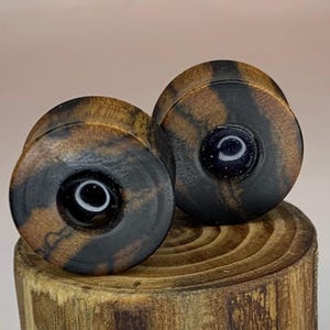 May include: A pair of wooden ear gauges with black and brown wood grain and a black and white inlay. The gauges have a round shape and are about 1 inch in diameter.