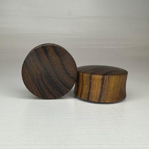 May include: Two wooden ear gauges, one is a flat disc and the other is a cylinder. Both are dark brown with a wood grain pattern.
