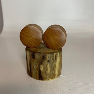 May include: Two round, wooden plugs with a cross-hatched design sit atop a small, cylindrical piece of wood. The plugs are a light brown colour, and the wood they rest on has a darker, natural wood grain.